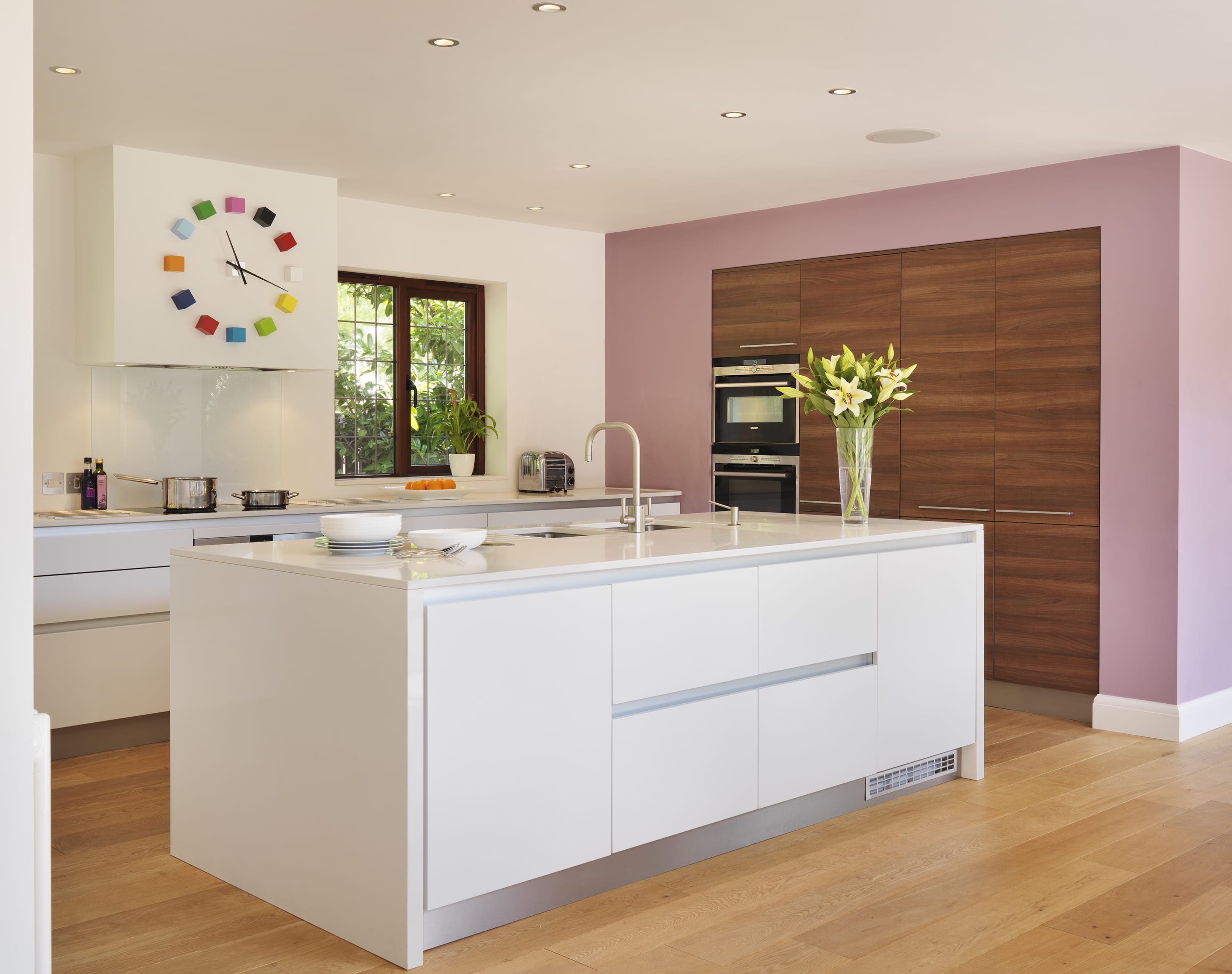 A modern fresh kitchen has a splattering of bright colour and pink feature wall. The look is kept uncluttered with the addition of recessed spot lights .