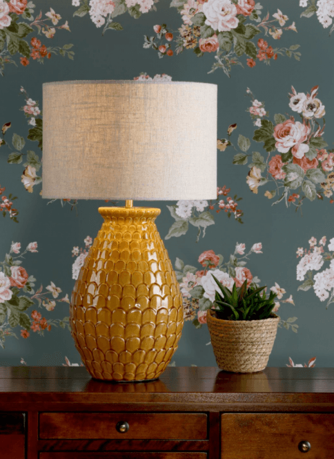Orange table lamp in front of green floral wallpaper.