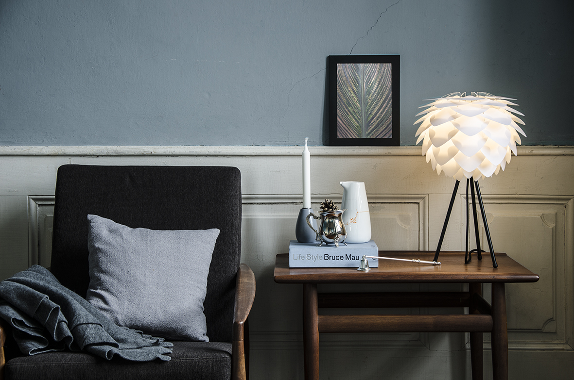 an arm chair next to a desk with a table lamp with a candle and books