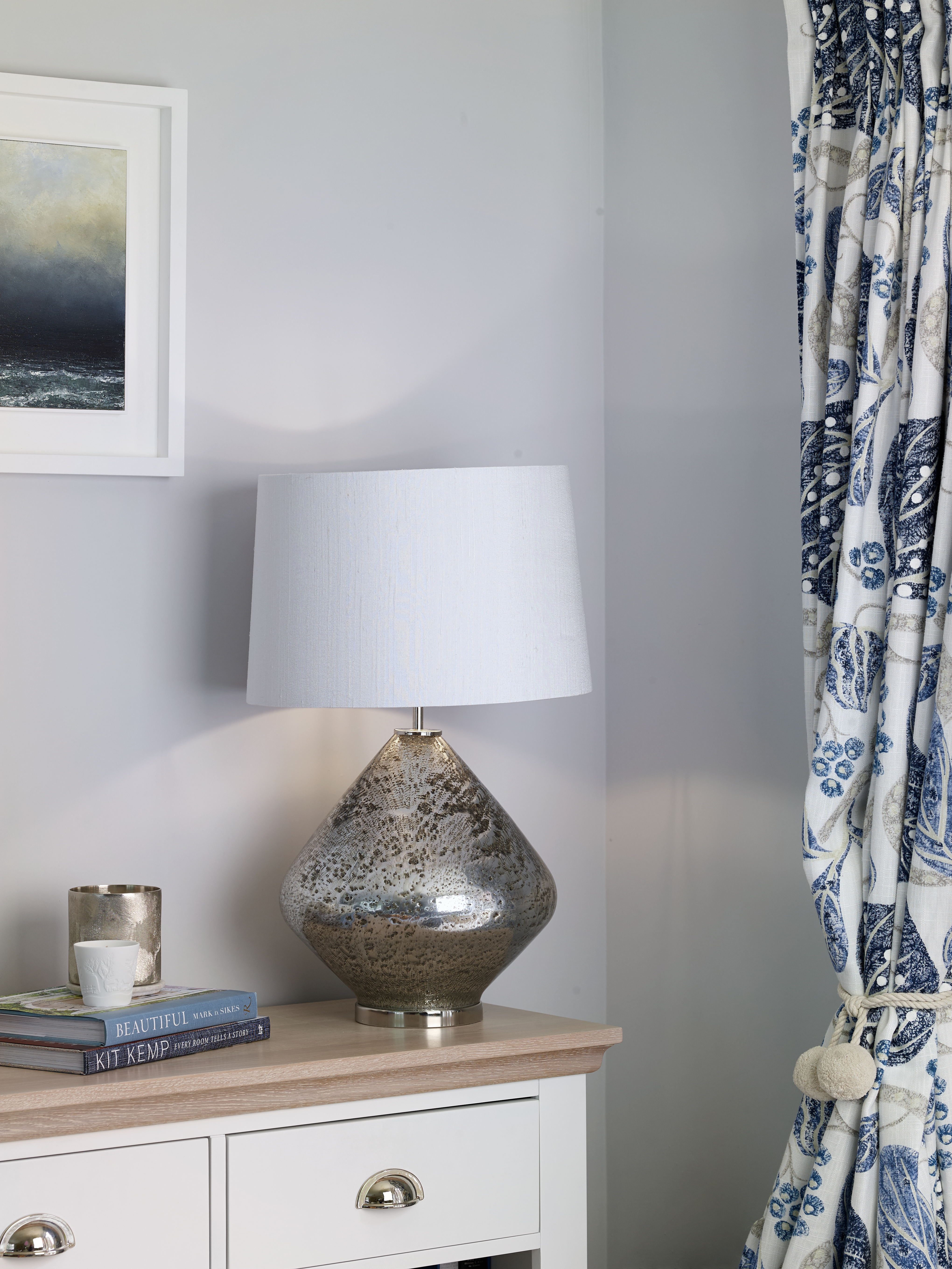 A table lamp with a stack of books and candles. Next to it is displayed blue patterned curtains with a beach themed pictured behind.