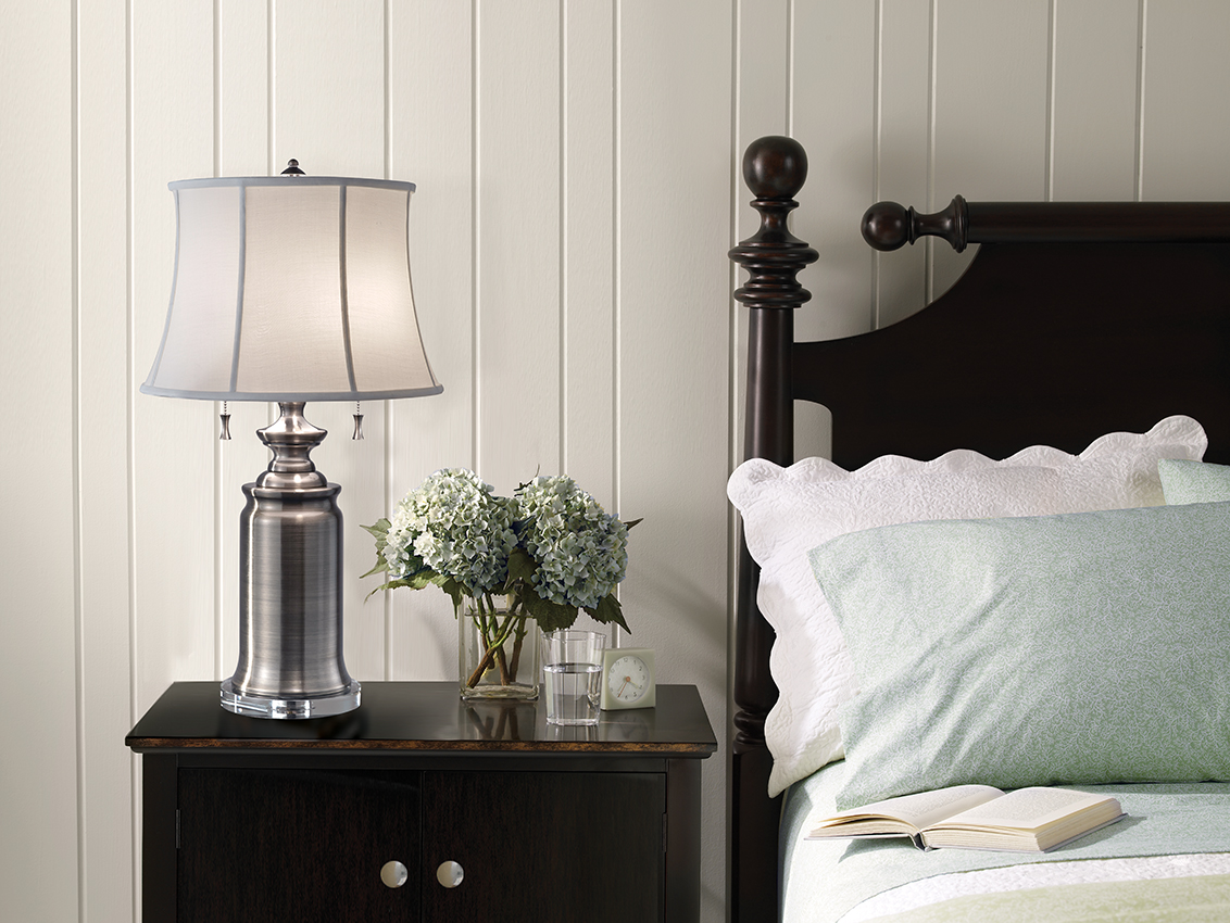 A table lamp on a bedside cabinet with a vase of flowers, cup of water and clock. Placed next to a bed with a book laid open.