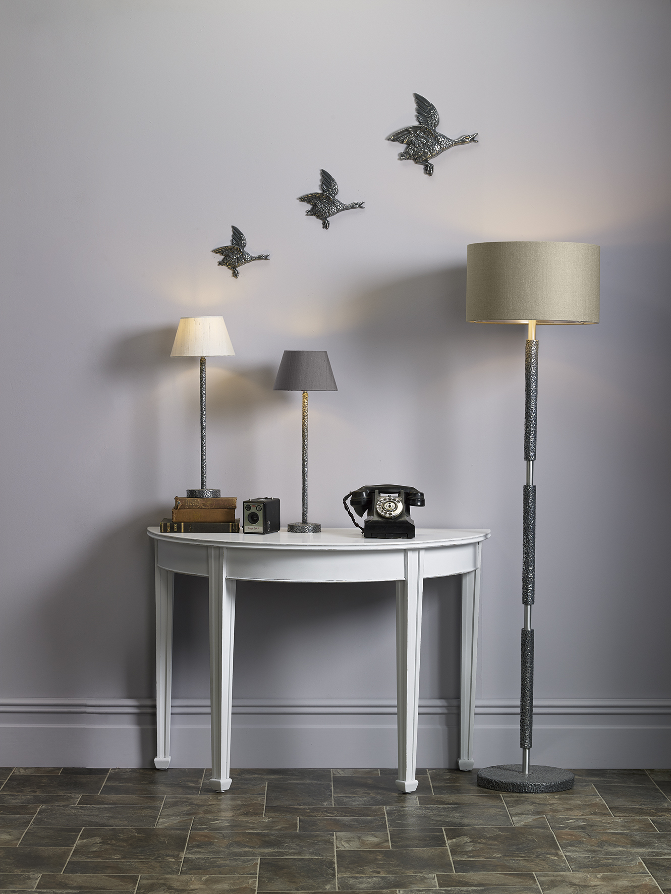 A floor lamp situated next to a side table with two table lamps with an old fashioned black phone. 3 birds are placed on the wall behind.