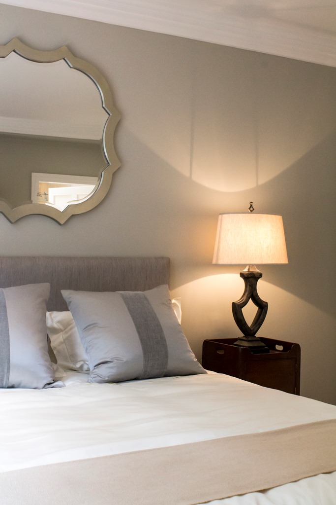 A bedroom with a large mirror hanging over a bed with a table lamp next to it.