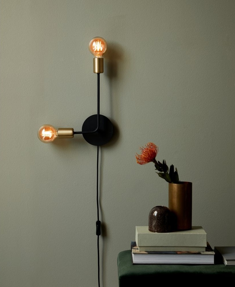 L shaped wall light on a green wall next to a stack of books. A pot plant is placed over the book.
