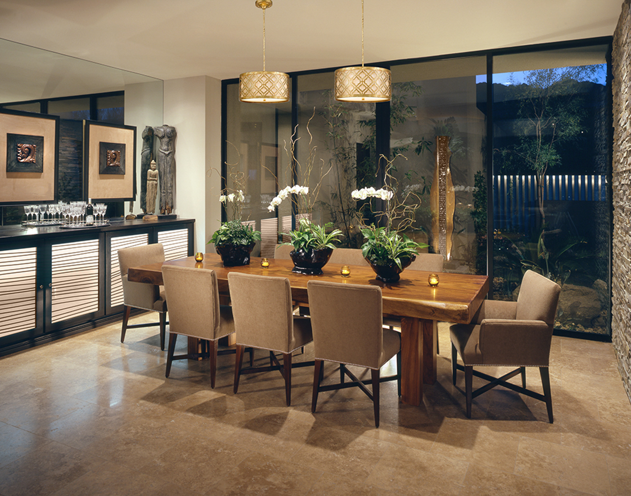 home dining room with eight chairs, hanging gold ceiling lights above glossed wood table. white and green flowers on table in modern setting