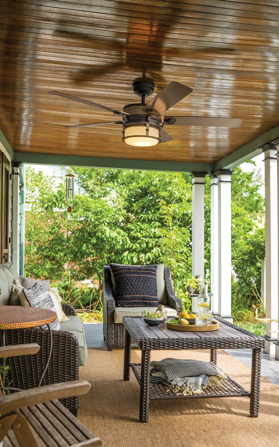 Out door setting with large green treem two rattan gry chairs and matching table. ceiling fan light on patio.