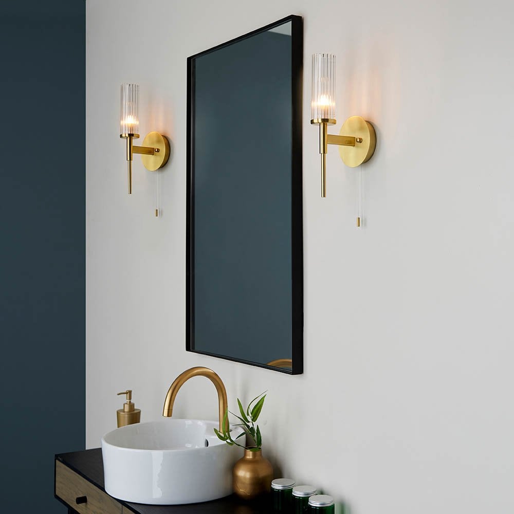 a modern bathroom with large black mirror with gold and ribbed glass wall sconces either side