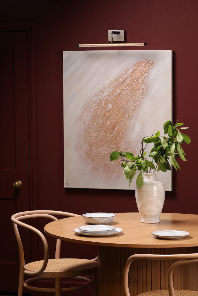 a modern slimline picture light above a piece of art on a purple wall with a green plant in a cream vase in front